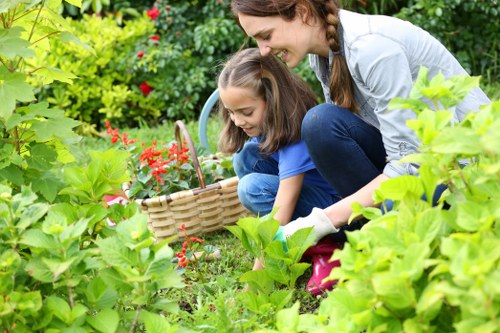 A woman and a young girl are gardening outdoors in a lush, well-maintained backyard, surrounded by vibrant green foliage, flowering plants, and shrubbery. The woman is crouched down, smiling and assisting the girl as they collect flowers or weeds into a wicker basket, which is held by the girl. The garden features a varied mix of dense plants, with some flowering bushes displaying red blossoms, and a background of trees and hedges providing privacy and a natural boundary. The grass lawn is neatly trimmed and dense, with a clear, flat paved patio area visible to the side. The scene is set in bright, natural daylight, suggesting a pleasant day suitable for outdoor gardening activities. The image reflects a well-kept outdoor space ideal for professional gardening and lawn care services, such as those offered by Lawn Mowing Ruislip, and showcases the natural and inviting environment of a typical suburban garden in the Ruislip area, potentially within the HA postcode district.