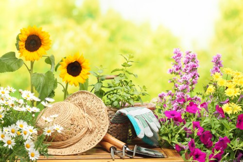 A vibrant garden scene featuring a wooden outdoor table set against a backdrop of lush green foliage. The table is decorated with a collection of gardening tools, including a pair of pruning shears, a small trowel, and a watering can, alongside a straw sun hat providing shade from the sun. Surrounding the table are flowering plants such as bright yellow sunflowers, clusters of purple and magenta flowers, and white daisies with yellow centers, creating a colourful and inviting atmosphere. In the background, blurred greenery and trees suggest a well-maintained outdoor space typical of gardens in Ruislip, with natural sunlight highlighting the textures of the leaves and blossoms. The scene conveys a peaceful, cultivated garden area suitable for outdoor gardening activities and lawn care, reflecting the services offered by Lawn Mowing Ruislip for local residential outdoor spaces in the HA4 postcode area.