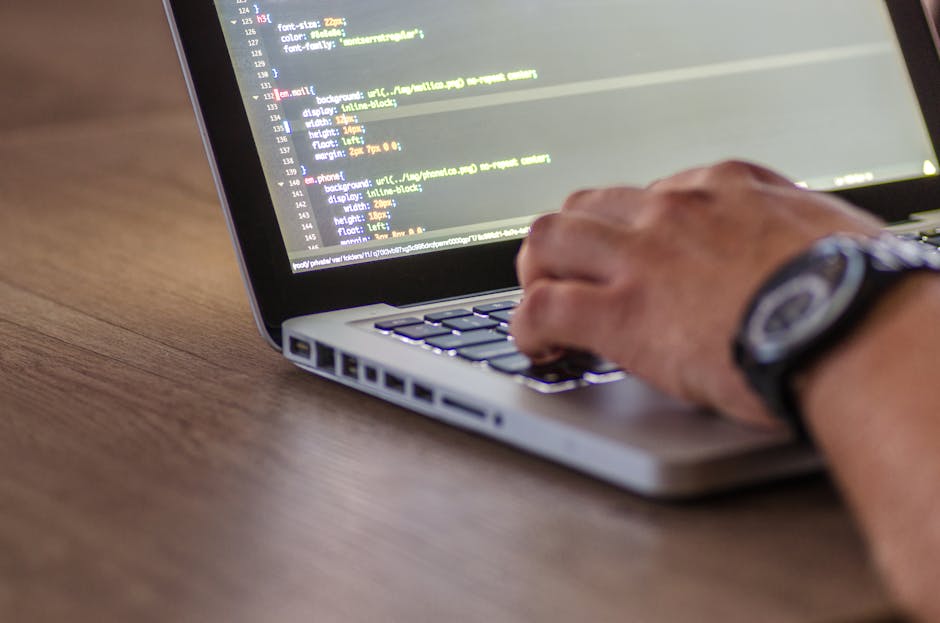 A close-up view of a person's hand operating a laptop computer on a wooden desk, with a focus on the screen displaying lines of code. The individual is wearing a wristwatch, and the environment sugges