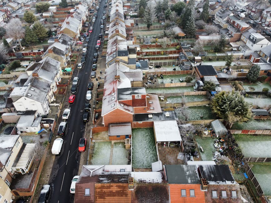 An aerial view of a suburban residential street showing rows of terraced houses with narrow front gardens, some with snow cover on rooftops and gardens. The street features parked cars alongside the p