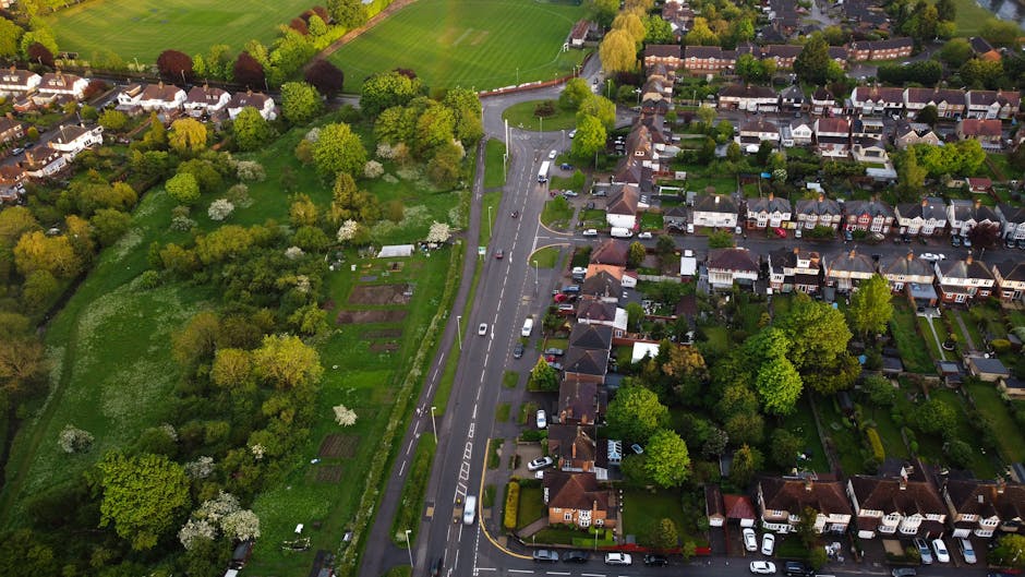 An aerial view of a suburban residential street showing rows of terraced houses with narrow front gardens, some with snow cover on rooftops and gardens. The street features parked cars alongside the p